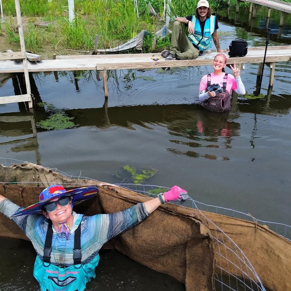 Trije nasmejani prostovoljci med delom na pomolu in v ozelenjenem vodnem bazenu.