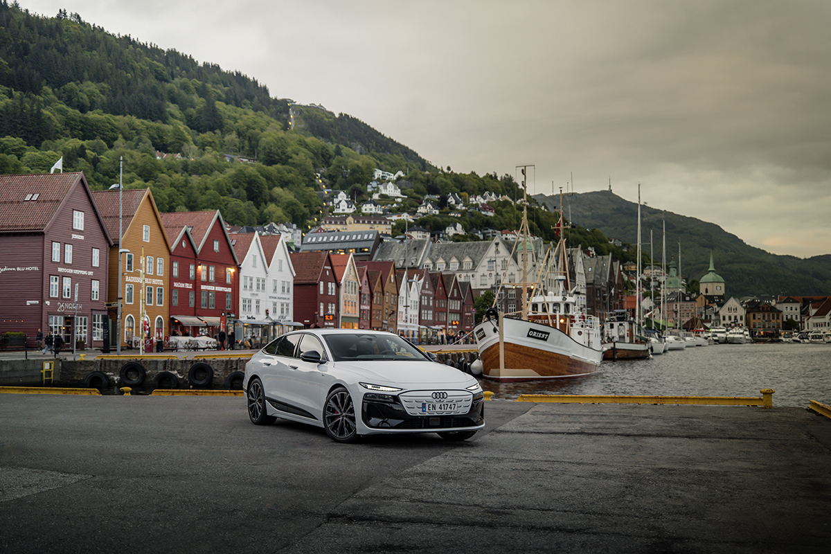 Bel Audi A6 Sportback e-tron parkiran pred ribiško ladjo in barvitimi hišami v ozadju.