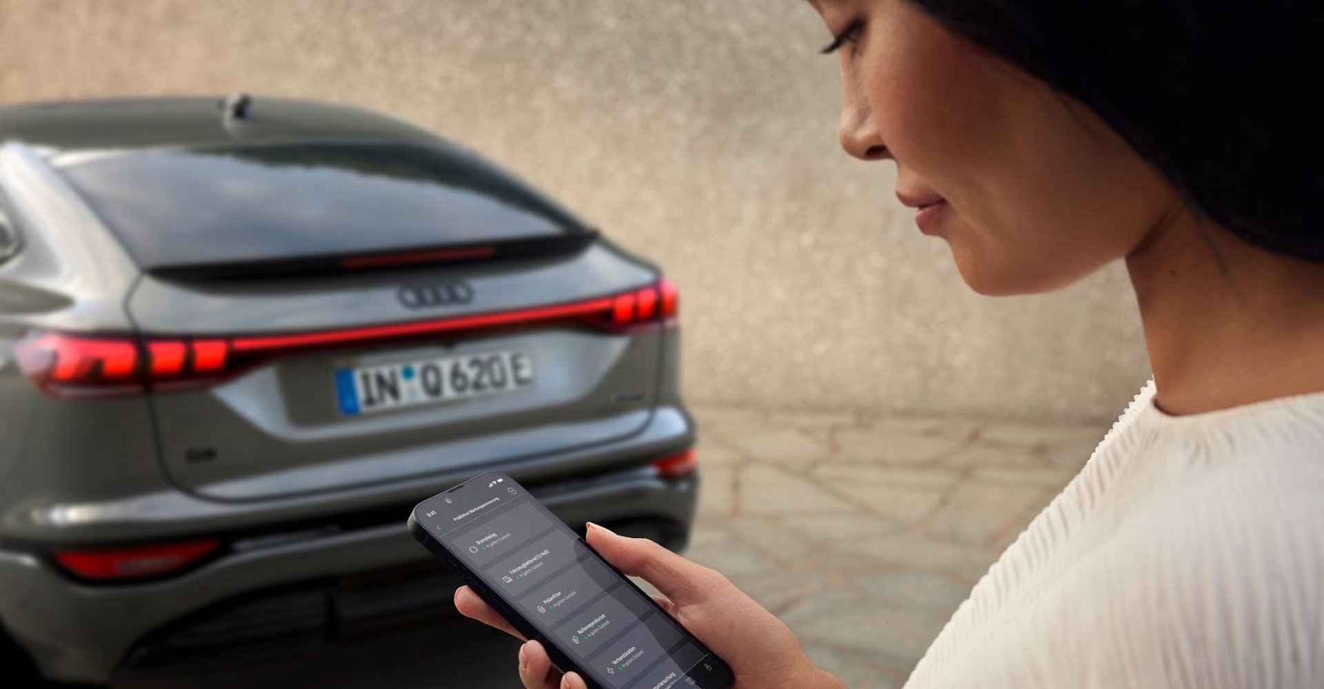 Ženska gleda na zaslon mobilnega telefona z aplikacijo myAudi. V ozadju je Audi Q6.