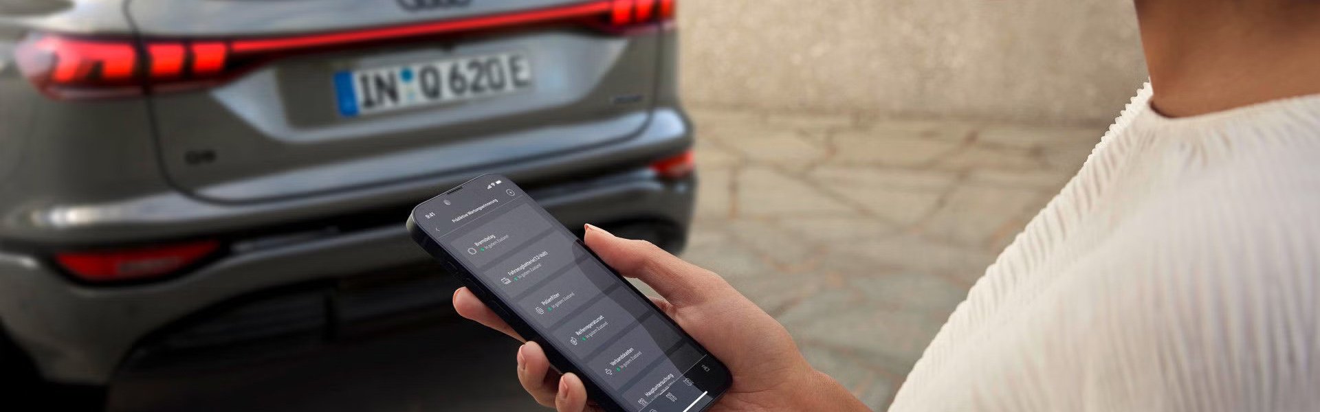 Ženska gleda na zaslon mobilnega telefona z aplikacijo myAudi. V ozadju je Audi Q6.