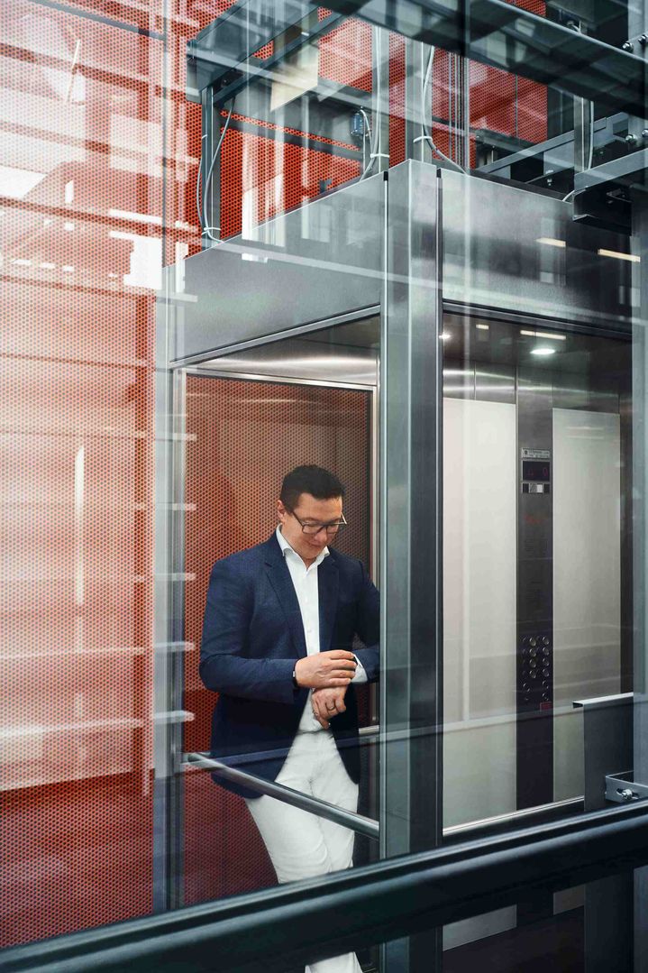 Giorgio Delucchi stoji v steklenem dvigalu in gleda na ročno uro. 