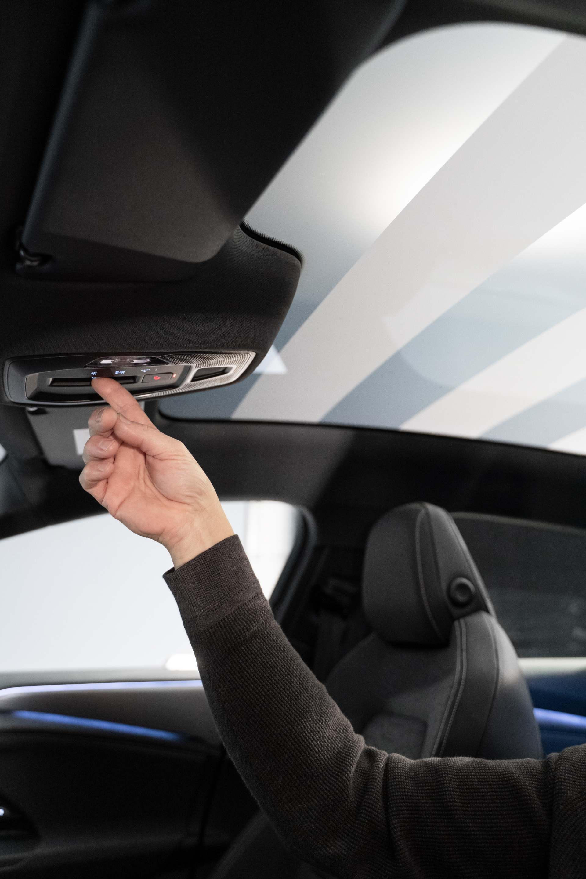 Pogled v notranjost vozila skozi voznikova vrata modela Audi A6 Sportback e-tron. Roka sega proti upravljalnemu elementu v stropni oblogi. Panoramska streha je zasenčena v progastem vzorcu.