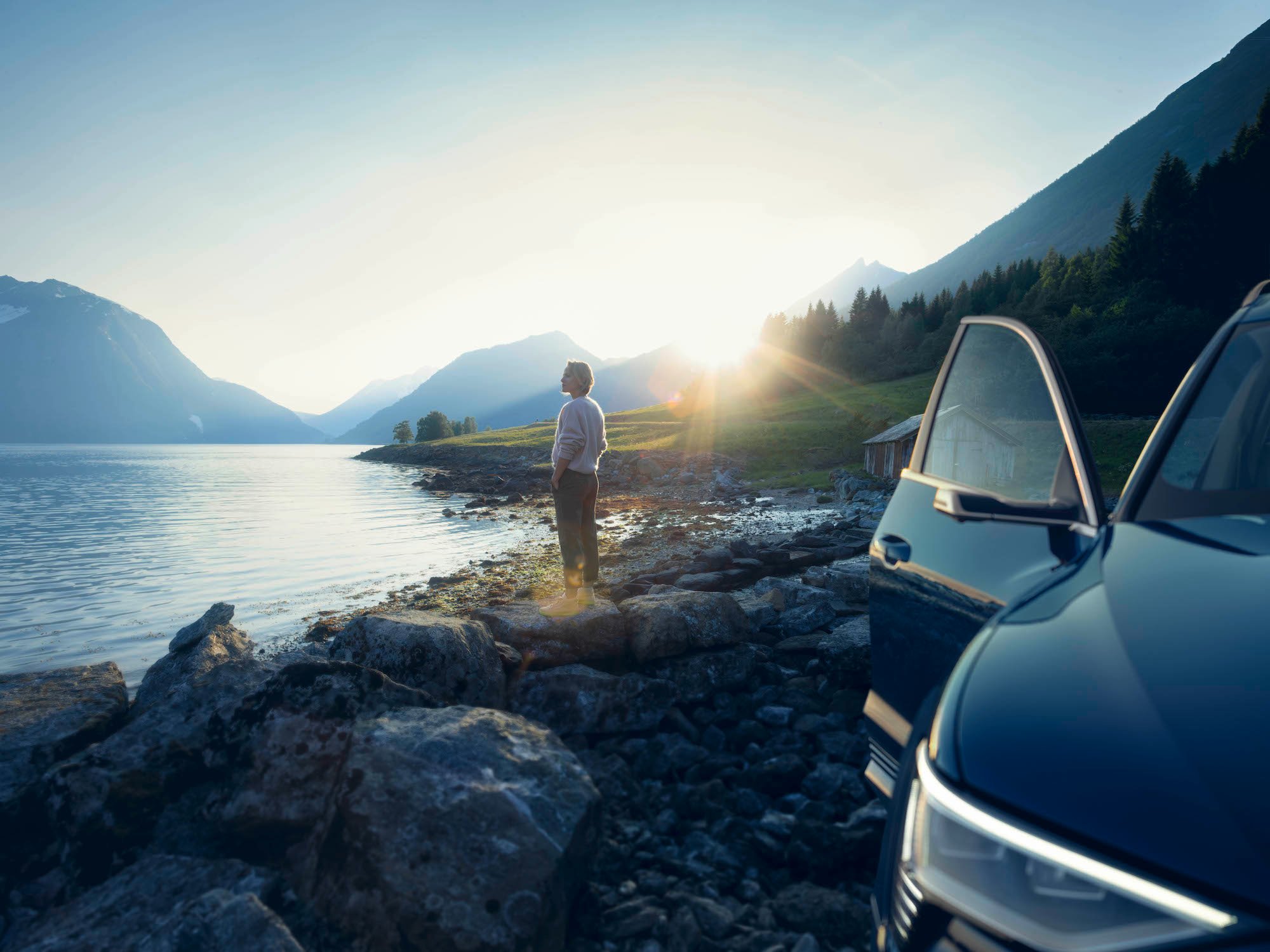 Audi terenec na obali jezera med sončnim vzhodom, v ozadju ženska, ki gleda na jezero