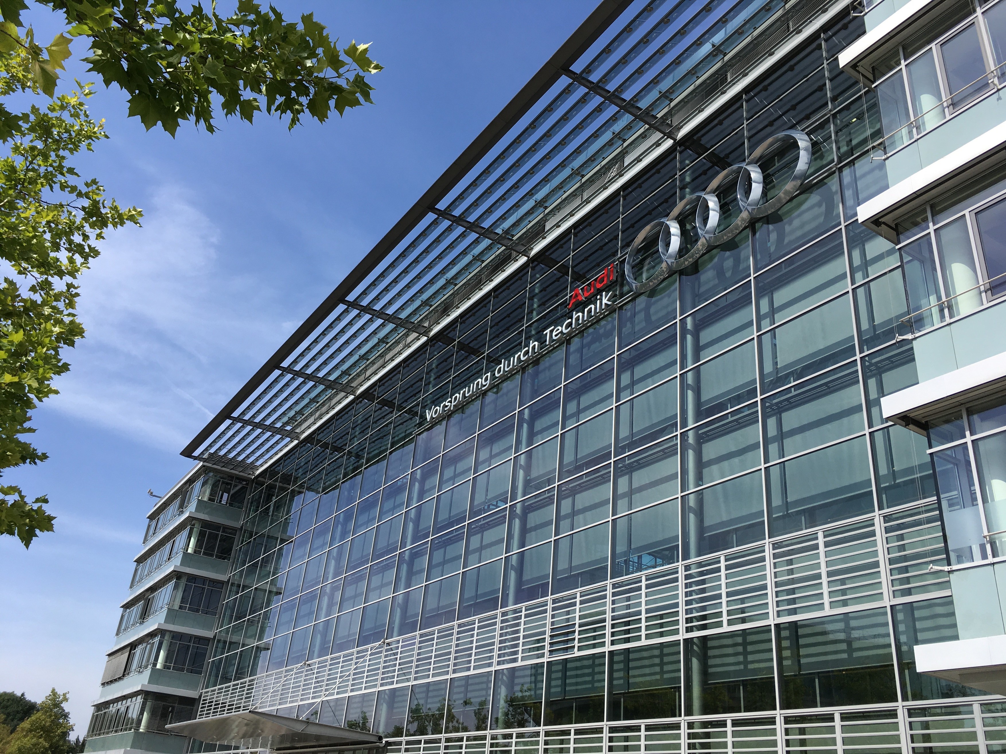 Steklena moderna zgradba z Audi logotipom, na levi strani drevo, nad zgradbo modro nebo.