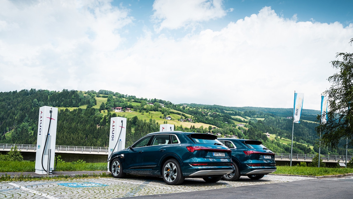 Dva modra vozila Audi e-tron na parkirišču na hribu. V ozadju zelen hrib in delno oblačno modro nebo.