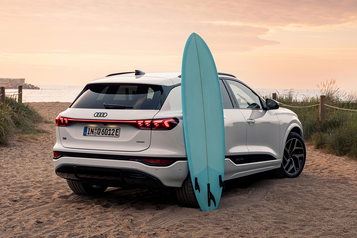 Bel Audi Q6 SUV e-tron je parkiran na peščeni plaži, v ozadju je morje. Na zadnjih vratih sloni turkizna deska za deskanje na vodi.