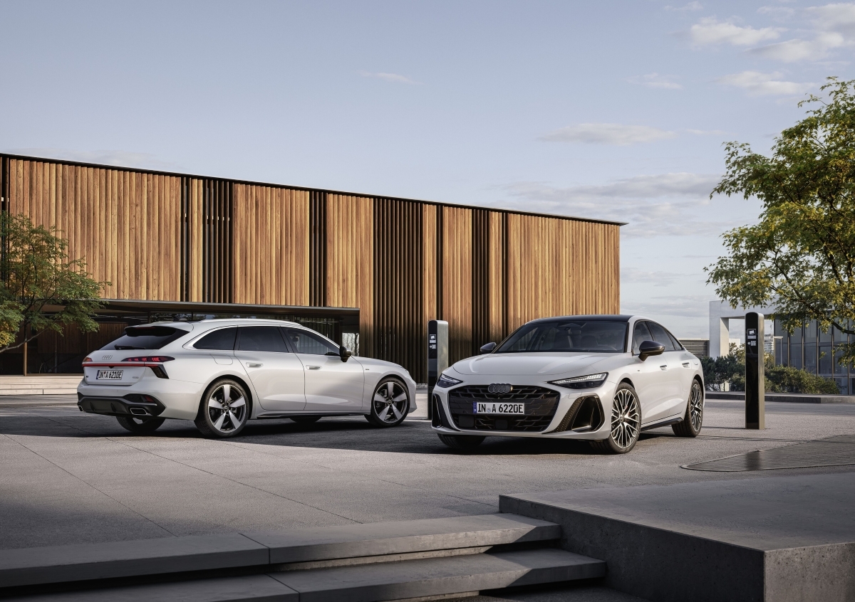 Beli Audi A6 Avant e‑hybrid quattro in beli Audi A6 Sedan e-hybrid quattro parkirana pred sodobno stavbo in priklopljena na polnilni postaji.