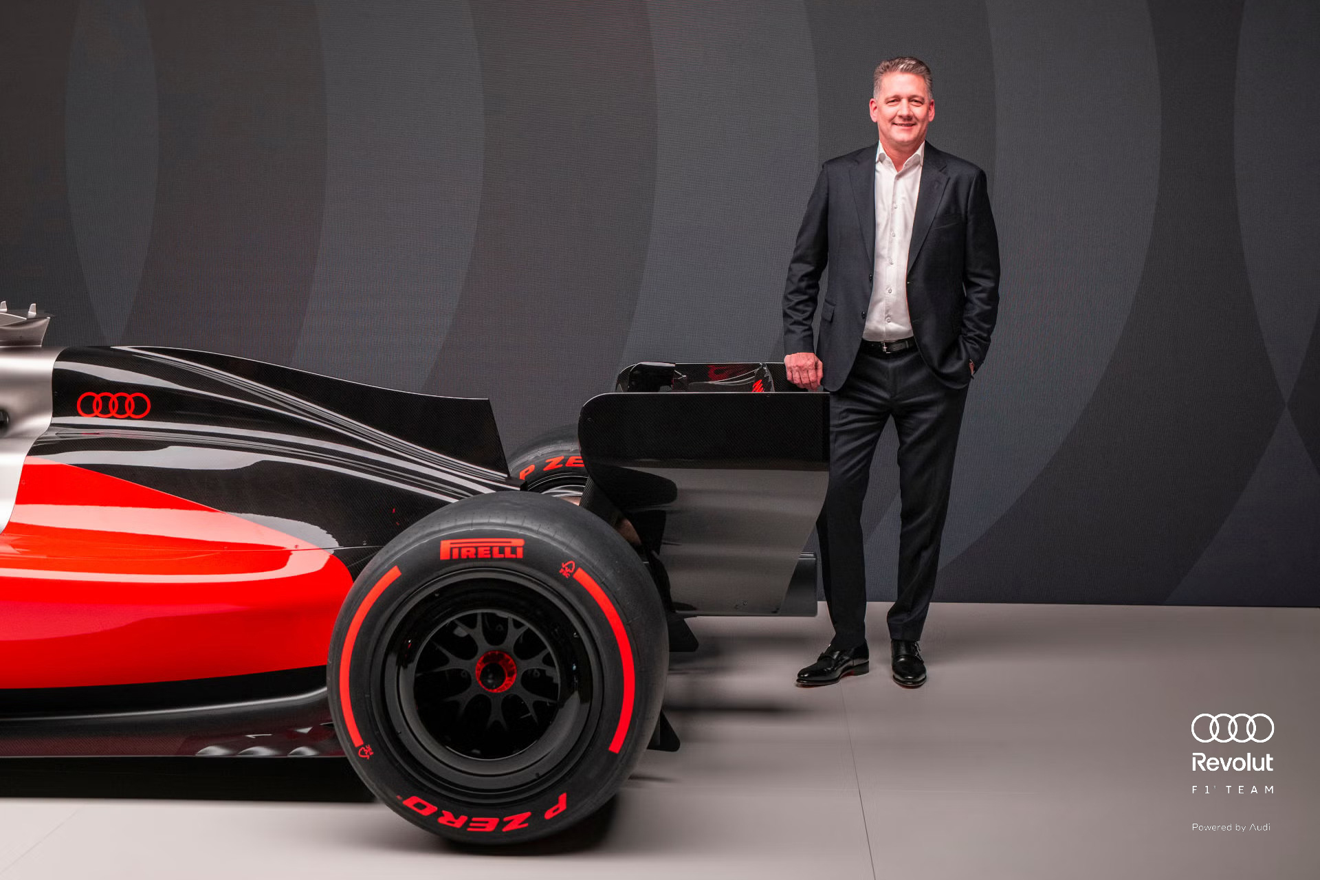 Dirkalnik Audi R26 za Formulo 1® v črno-sivo-rdeči barvni shemi v temnem studiu, na njem so vidni logotipi sponzorjev, ob njem pa stoji izvršni direktor družbe Audi Gernot Döllner s prekrižanimi rokami.