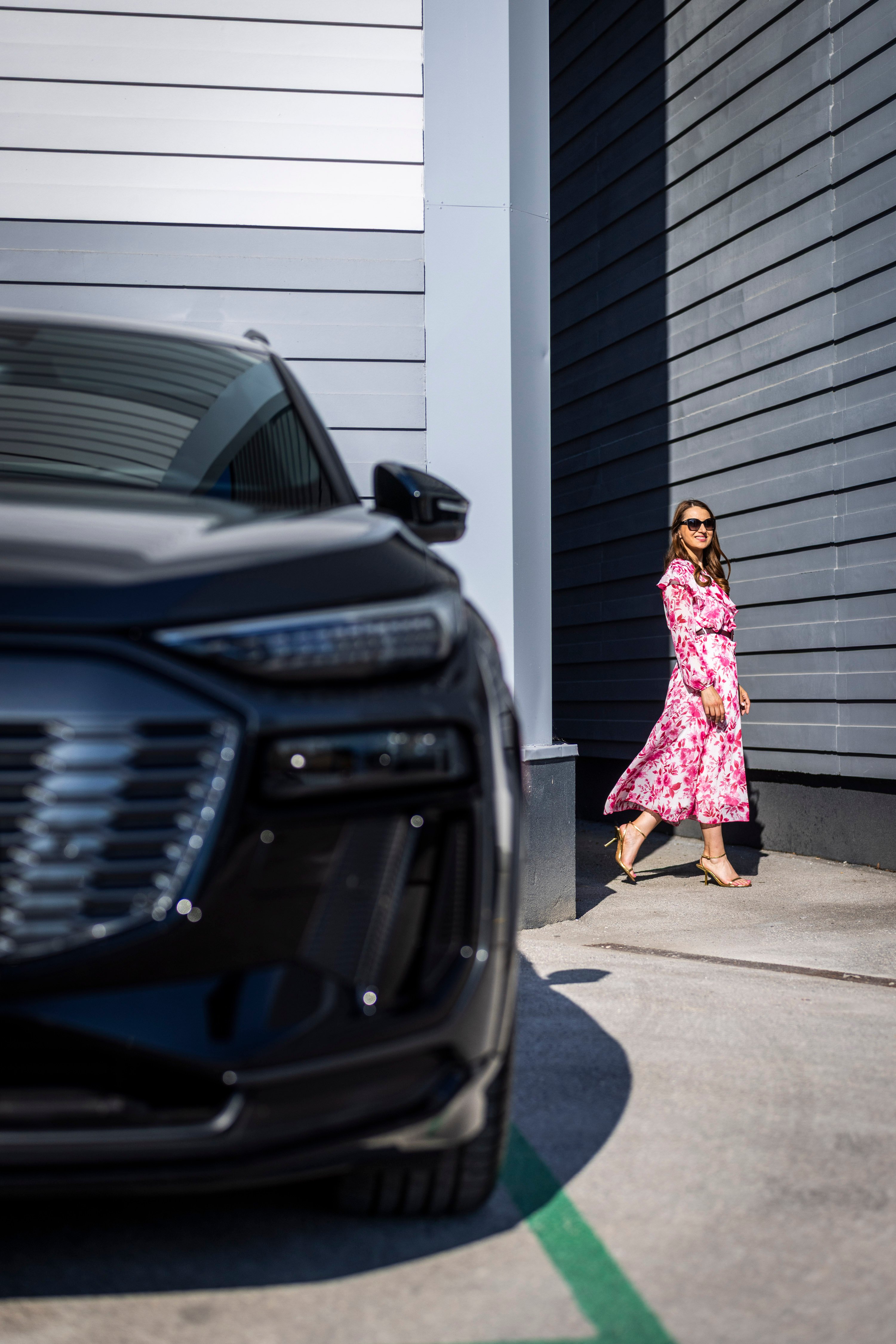Mirela Župančič hodi ob elegantnem Audiju Q6 e-tron pred moderno, sivo progasto stavbo.